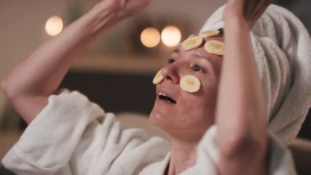 お風呂の後に手入れの行き届いた女性の閉じる彼女の肌の世話栄養と持ち上げ肌のために顔に皮をむいたバナナスライスを入れて — ストック動画