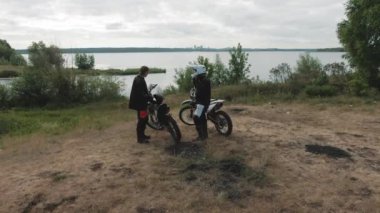 Motorsikletlerinin önünde duran iki adamın göl ya da göl kenarına park etmiş ve teçhizatlarını çıkarırken insansız hava aracı görüntüsü.