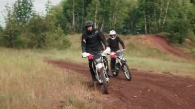 Ormanda toprak yolda motosiklet süren kasklı ve teçhizatlı iki adamın izini sürüyoruz.