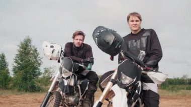Kasvetli bir günde motosiklet süren genç adamların kasvetli bir şekilde dışarıda oturup kameraya baktıkları orta boy bir çekim.