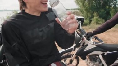 Motosiklete binen ve arkadaşlarıyla sohbet ederken su içen neşeli bir gencin orta boy fotoğrafı.