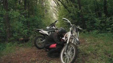 Miğferli adamların orta ölçekli görüntüleri ve motosikletin önünde çömelip aşırı gelişmiş ormandaki mekanik arızayı düzeltmeye çalışan binicilik takımları.