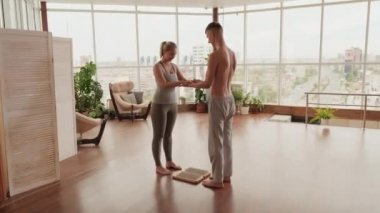 Erkek yoga koçu, beyaz bir kadının yoga sınıfında panoramik pencereli çivilerle sadhu tahtasında durmasına yardım ediyor.