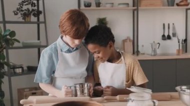 Mutfaktaki masada beyaz önlük giymiş, yukarı bakıp kameraya gülümseyen iki çoklu etnik okul çocuğunun görüntüsü. Kızıl saçlı Kafkasyalı un süzgeci, Afrikalı arkadaşı onu izliyor.
