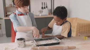 Kızıl saçlı, beyaz önlüklü Afrikalı ve beyaz okul çocukları mutfaktaki masada duruyor, kurabiye serpiştiriyorlar.