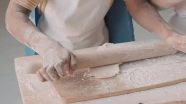 Yüksek açılı, önlüklü, elleri un kaplı, tahta kesme tahtasına çiğ hamur işi serpiştirilmiş, tanınmayan birinin yakın plan fotoğrafı.