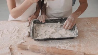 Mutfak masasının yanında durup un döken, kalp ve yıldız şeklinde kurabiyelerle fırın tepsisine un döken iki kişinin yüksek açılı görüntüsü.