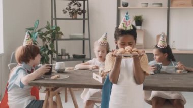 Orta boy Afrikalı okul çocuğu önlük giyiyor ve parti şapkası dolusu kurabiye taşıyor, onları kokluyor, kameraya gülümsüyor, mutfak masasının önünde duruyor, çeşitli arkadaşlar yemek yiyor ve konuşuyor.