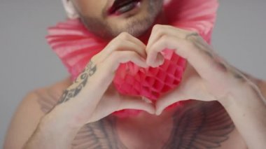 Yakın çekimde, sırtında beyaz saçlar, boynunda parıltı ve kırmızı bir fırça olan dövmeli bir adamın el hareketi ve kameraya üfleyerek öpüşmesi var.