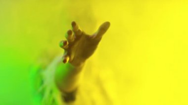 Dramatik makyajlı ve şık ceketli bir adamın portresi. Yeşil ve sarı ışıkta dalgalanan dumandan eline ulaşıyor.