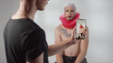 Elinde cep telefonu olan bir adamın orta boy fotoğrafı ve boynunda dramatik bir makyaj ve kırmızı yaka olan üstsüz bir erkek modelin fotoğrafını çekiyor.