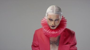 Dramatik makyajlı, parıltılı beyaz saçlı, boynunda kırmızı yaka ve göğsünde kamera önünde poz veren bir adamın portresi.