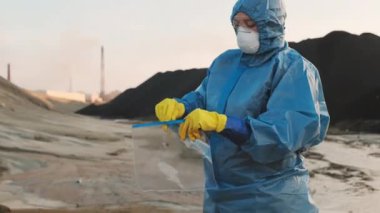 Zehirli tehlikeli bölgede koruyucu giysi üzerine ekoloji uzmanının orta ölçekli görüntüsü kirlenmiş toprakla birlikte özel bir paketin içine test tüpü yerleştiriyor.