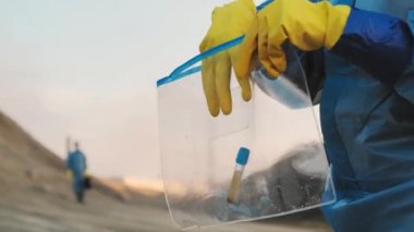Sarı eldivenli tanımlanamayan ellerin yakın çekimleri kirlenmiş topraktan özel şeffaf plastik torbaya test tüpü yerleştiriyor.
