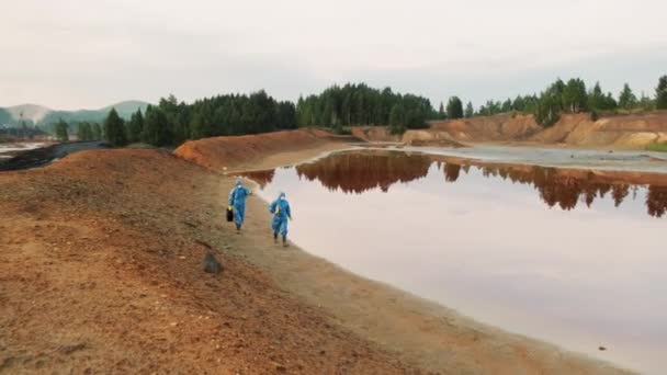 两名身穿蓝色防护服的女性生态专家在污染最严重的地区之一的池塘边与肮脏的有毒水一起行走时被拍的高角镜头 — 图库视频影像