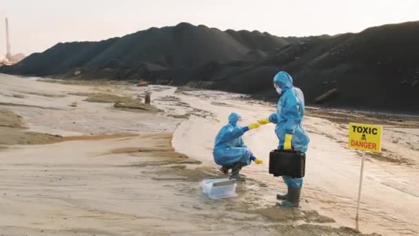 汚染された水や土壌の品質管理テストのためのデータを収集し 背景にほこりや廃棄物の黒い丘を持つ保護衣装の女性同僚の完全なショット — ストック動画