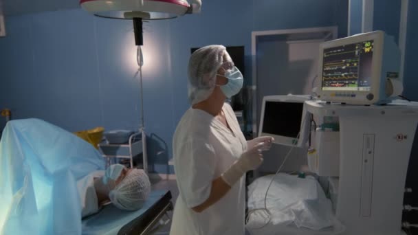 ナルコシス下の手術台に横たわっ患者の重要な機能を示すディスプレイを調整白いスクラブで外科アシスタントの中ショット — ストック動画