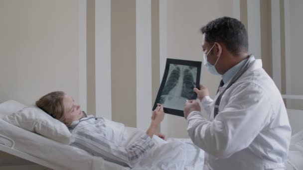 合资格男医生的侧视中照 向躺在卧床内解释详情的年轻女子展示胸部X光检查结果 — 图库视频影像