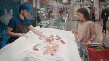 Alışveriş arabalı mutlu bir genç kadın, erkek satıcısıyla konuşurken büyük süpermarkette günlük alışverişte taze kırmızı balığı seçerken gülümsüyor.
