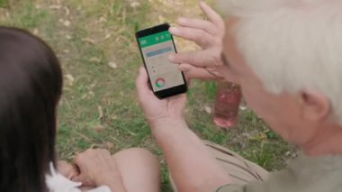 Beyaz saçlı beyaz adamın ve karısının arka planda çimenlikte yan yana oturup cep telefonu ekranına bakmaları ve konuşmaları.