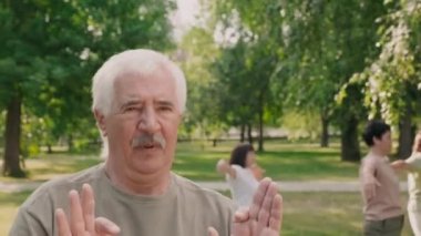 Açık havada Qigong antrenmanı yapan, bıyıklı, neşeli, beyaz, yaşlı bir sporcunun orta boy portresi.