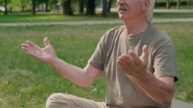 Beyaz saçlı ve bıyıklı yaşlı beyaz bir adam parktaki mindere oturmuş yoga yapıyor.
