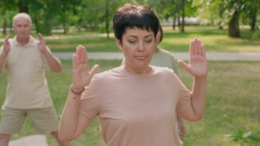 Farklı yaşlarda beş aktif yetişkin insan parkta çim üzerinde yoga minderleri üzerinde durup denge egzersizi yapıyor.