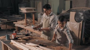 Küçük çocuğun kareli gömlekli ve önlüklü halinin orta büyüklükte görüntüleri. Babasının talimatları doğrultusunda marangozluk projesi için ahşap korkulukları yığıyor.