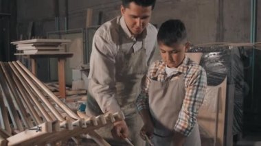 Baba ve oğlunun marangozluk atölyesinde çalı çırpı döşeyen ahşap iskemlenin yapımını bitirdiği orta boy bir fotoğraf.