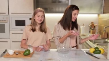 Neşeli, çekici bir kadın ve genç kızının parlak, modern mutfakta yemek pişirirken avokado keserken ve kavanozda krema karıştırırken kamerayla konuşurken orta boy fotoğrafı.