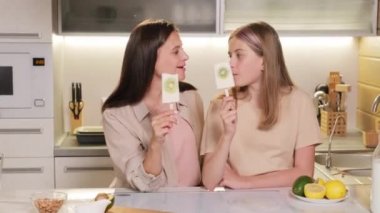 Medium slow-motion portrait of joyful mother with pretty teenage daughter looking at camera smiling while eating homemade fruity ice-cream on stick