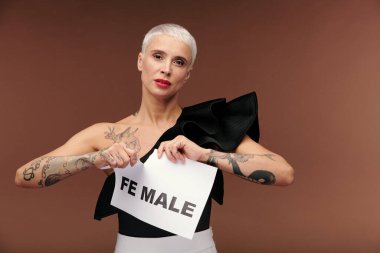 Siyah tanktop giymiş sarışın zarif bir kadın kahverengi arka planda kameranın önünde dikilirken hece ve kelime erkeğiyle yırtılıyor.