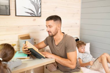 Küçük kızına açık kitap okuyan genç bir baba. Arkasında dokunmatik ped kullanan sevimli bir oğlanla aynı masada oturuyor.