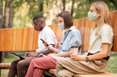 Koruyucu maskeli genç esmer kadın akıllı telefondan kayarken dizüstü bilgisayarı olan Afrikalı bir adamla içkili bir kız arasında oturuyor.