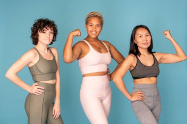 Eşofmanlı, çeşitli etnik kökenlere sahip üç genç kadın kameranın önünde sıraya girip mavi arka plana karşı egzersiz yapıyor.