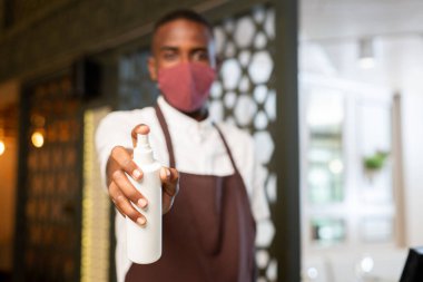 Üniformalı ve koruyucu maskeli Afrika kökenli genç garsonun eli, restoran ya da kafe müşterilerine sprey sıkacak.