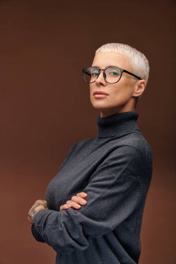 Koyu gri kazaklı, gözlüklü genç bir kadın kameranın önünde dikilirken kollarını göğsünden kavuşturuyor.