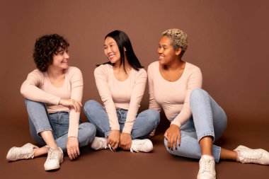Group of young casual females of various ethnicities sitting on the floor against brown background, looking at each other and chatting clipart