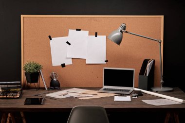 Ofis malzemelerinin bulunduğu, mobil cihazlar, planlar, lamba ve sepetin bulunduğu karton panoların olduğu bir mühendislik işyeri.