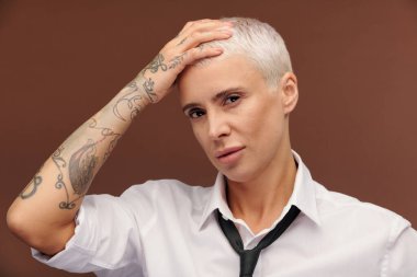 Kolunun her tarafında dövmeleri olan orta yaşlı bir iş kadını ellerini başının üstüne koyarak sana kahverengi arka planda bakıyor.