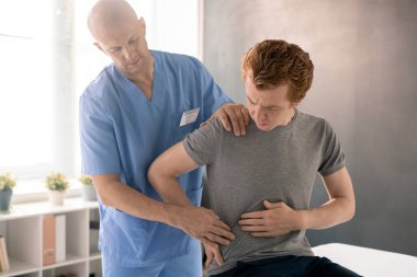 Hasta adam, klinikteki rehabilitasyon eğitiminde tıbbi kanepede otururken sırtına dokunmanın ve yaralanmanın yerini gösteriyor.