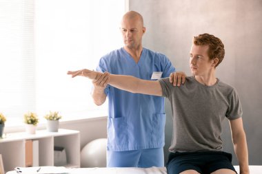 Rehabilitasyon kliniklerinde egzersiz yaparken kanepede oturan genç bir hastayı destekleyen üniformalı kel ve olgun bir fizyoterapist.