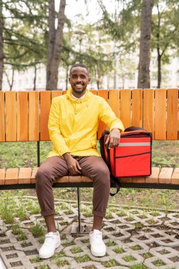 Üniformalı genç erkek kurye, parkta bankta oturup dinlenirken müşterilerin siparişleriyle birlikte büyük kırmızı sırt çantasını tutuyor.