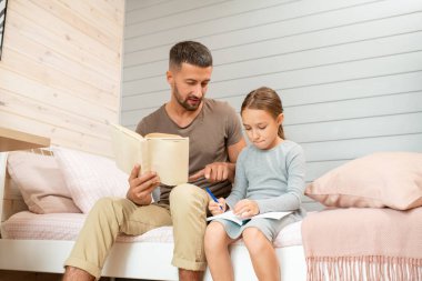 Küçük tatlı kızının notlarını gösteren açık bir kitabı olan genç bir baba. Hem yatakta oturuyor hem de ev ödevlerini yapıyor.