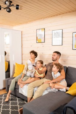 Genç, çağdaş bir çift ve gündelik giysiler içindeki iki tatlı çocukları oturma odasında kanepede oturup hafta sonları televizyon izliyorlar.