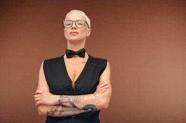 Siyah yelekli, yelekli, papyonlu, kollarını göğsünde dövmelerle bağlayan olgun sarışın bir kadın.