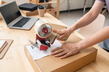 Çevrimiçi mağazanın kadın yöneticisinin elleri seloteyp silah dağıtıcısıyla karton kutuyu mühürlüyor ve müşterisi iş yerinin yanında paketleniyor.