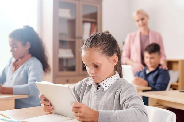 真剣にかわいいです女の子と彼女のクラスメートとともにデジタルタブレット座っています机と作業個別にオンラインテキストながら教師コンサルティングそれら — ストック写真