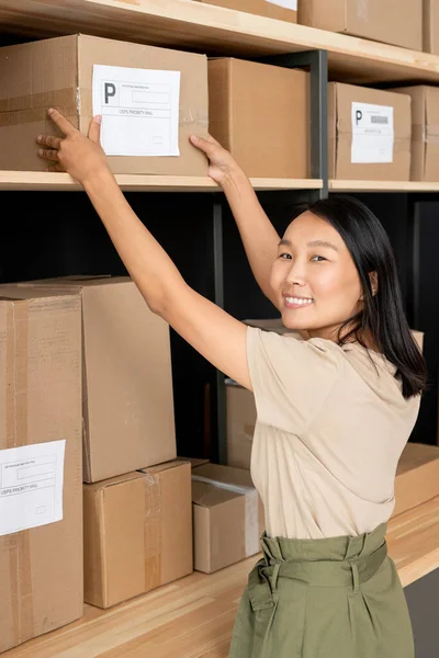 Genç, mutlu, esmer bir kadın yönetici büyük raftan müşterilerin siparişleriyle dolu kutulardan birini alıyor ve dişlek gülümsemeyle sana bakıyor.