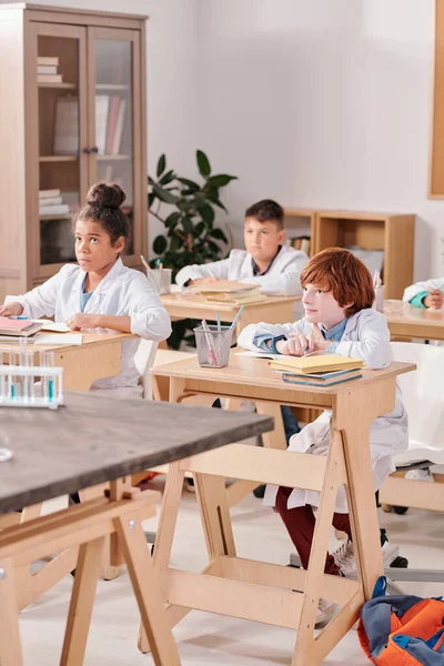 化学の授業で2列に机に座って先生の説明を聞いている白塗りの小学生のグループ — ストック写真